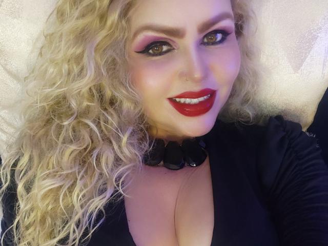 AnnaRichie with curly blonde hair, red lipstick, black choker, and black top, showing cleavage, smiling at the camera with purple eye makeup. Her nose is pierced.
