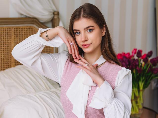 EricaDiaz with pink sweater, white shirt, and long brown hair, touching her face with a ring on her finger, looking cute and sexy, standing by a bed with flowers.