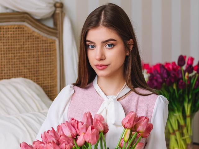 EricaDiaz with pink tulips, holding a bouquet, wearing a pink vest over a white shirt, brown hair, blue eyes, and a subtle smile, standing in front of a bed.