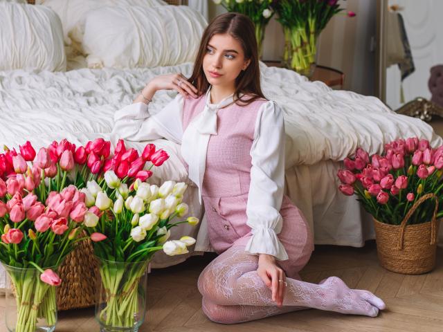 EricaDiaz with pink vest, white blouse, and lace tights, kneeling by bed, surrounded by pink, white, and red tulips in glass vases and a wicker basket.