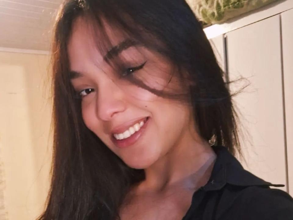 FeiticeiraX has long black hair, brown eyes, and is smiling with her tongue slightly out, wearing a black shirt, showing a small mole on her left cheek.