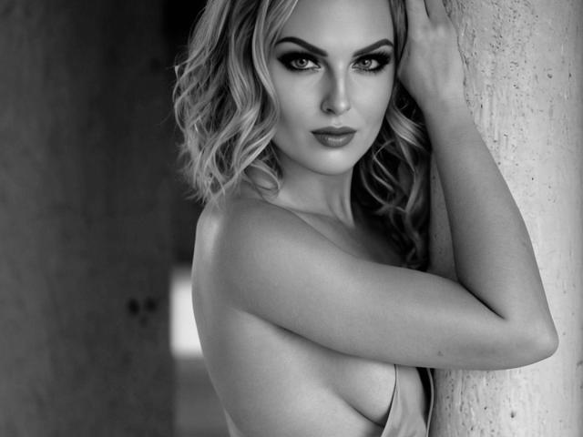 MeridianSexy with wavy blonde hair, sultry eyes, and full lips, leaning against a textured wall, topless, showing small, perky tits, with a sexy, confident expression. Black and white.