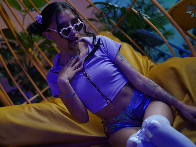 EviieGothel with heart-shaped sunglasses, purple crop top, shiny silver shorts, white thigh-high socks, and tattoos, sitting on a yellow chair, hand on chest, blue and yellow lighting.