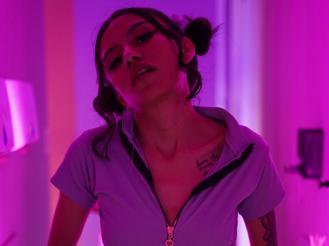 EviieGothel con cabello oscuro recogido en moños, iluminada en tonos morados, vistiendo una camisa con cremallera que deja ver un tatuaje en el pecho y otro en el brazo derecho, mirando hacia abajo de forma seductora..