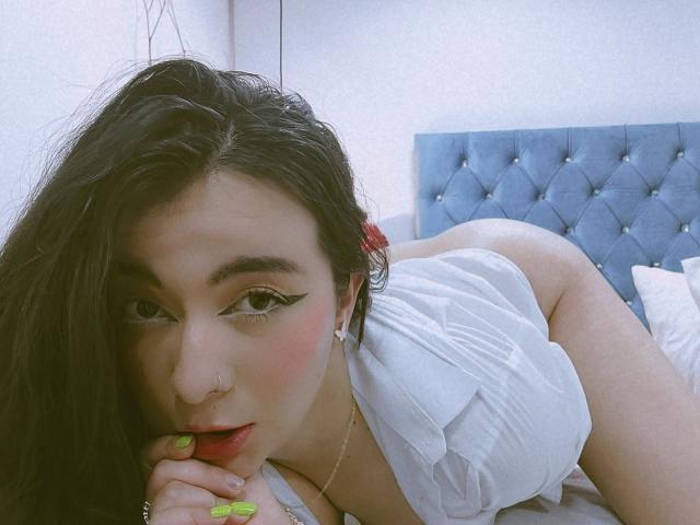 AnaGerald, avec ses longs cheveux noirs, sa chemise blanche et ses ongles verts, est allongée sur un lit, regardant la caméra. Elle a le nez percé, un tête de lit bleu avec des motifs, et montre ses fesses, avec les lèvres légèrement entrouvertes..