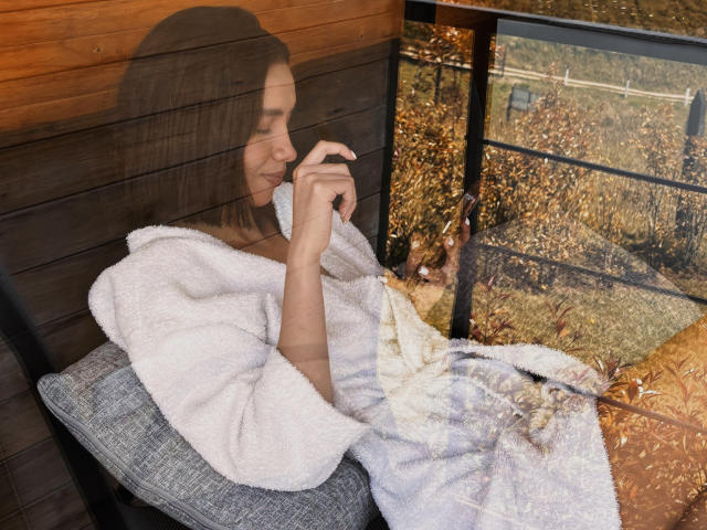 BonnieShyy, with long brown hair, sits on a gray pillow, wearing a white fluffy robe, her small tits visible, holding a phone, reflection on wooden wall.