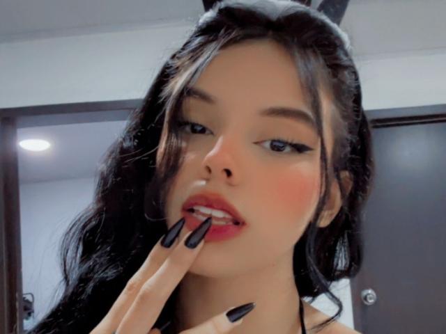 GuadalupeClak, con cabello negro largo, delineador negro y lápiz labial rojo, mordiéndose el dedo con uñas negras y puntiagudas, tiene una expresión sexy y sensual, y lleva una diadema negra..