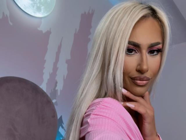 KarlaBart with long blonde hair, pink eyeliner, and pink lipstick, wearing a pink top, touching her chin, showing off her pouty ass-kissing lips.