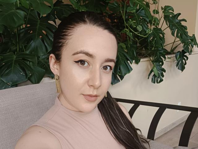 MabyLegrand with pale skin, brown eyes, and black hair in a ponytail, wearing a pink top, looking at the camera with a slight smile, sitting in front of green leaves.