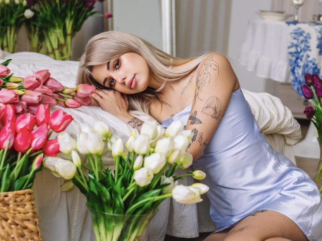 LaraMoss with platinum blonde hair, tattoos, wearing a silky blue dress, leaning on a bed with pink and white tulips, looking seductively at the camera.