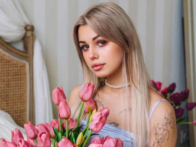LaraMoss with blonde hair, pink tulips, tattoos on arms, wearing a choker and light blue dress, holding flowers, looking at camera with a soft, seductive expression.