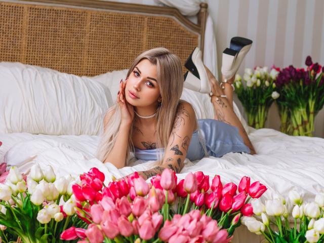 LaraMoss with long blonde hair, tattoos on arm, lying on bed, head on hand, wearing blue dress, white heels, surrounded by pink and white tulips, looking at camera.