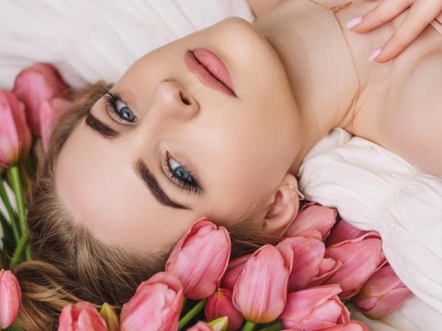 EvaDaly with blue eyes, pink lips, and blonde hair, lying on pink tulips, wearing a white shirt, and a gold necklace. Her tits are slightly visible.