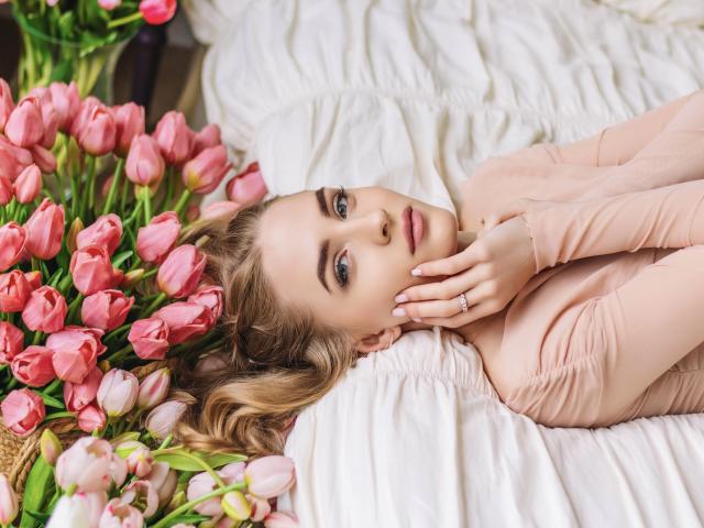 EvaDaly lies on a bed with pink tulips, wearing a pink long-sleeve top, her hand touching her face, looking at the camera with blue eyes and blonde hair.