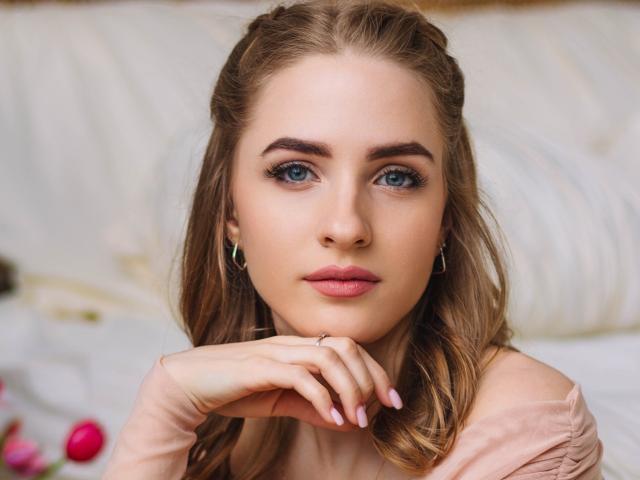 EvaDaly has blue eyes, light brown wavy hair, pink lips, and is wearing a pink top with her hand resting on her chin, showing off her pink nails.