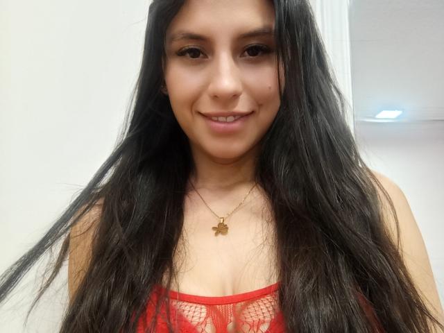 MadissonFontaine with long black hair, smiling, wearing a red lace top that shows her tits, gold necklace with a butterfly pendant, light skin, brown eyes, and a cute face.