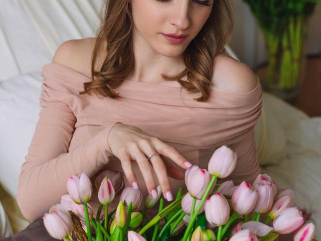 EvaDaly with pink off-shoulder top, gentle expression, light brown wavy hair, touching pink tulips with green stems, wearing ring on right hand, soft pink nails.