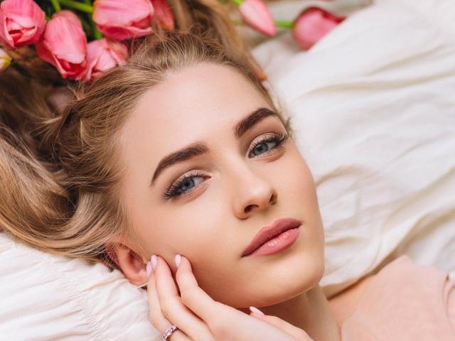 EvaDaly's face with pink tulips in her blonde hair, lying on white pillows, touching her cheek with pink-nail-polished fingers, blue eyes, and pink lips.