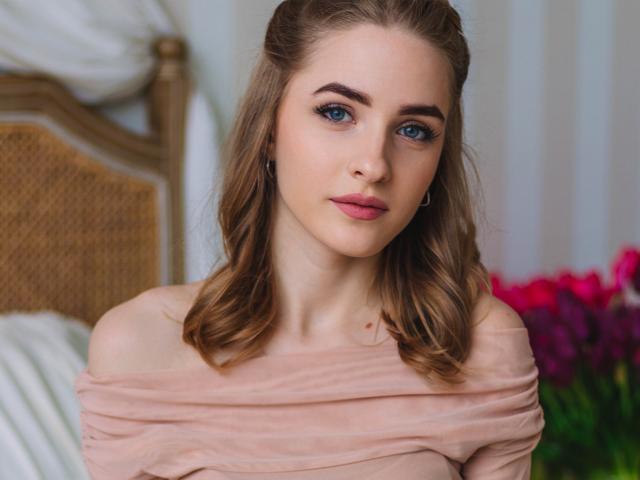 EvaDaly with blue eyes, wavy brown hair, pink off-shoulder top, slight smile, subtle makeup, small nose ring, standing in front of a wicker headboard with pink flowers.