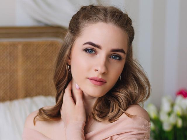 Eva Daly tem olhos azuis, cabelo loiro ondulado e batom rosa, vestindo uma blusa rosa com um ombro de fora e uma mão no pescoço, olhando diretamente para a câmera..