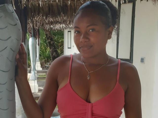 MiaTight with brown skin, black hair in a bun, wearing a pink top showing her big tits, silver necklace, standing next to a white column, smiling.