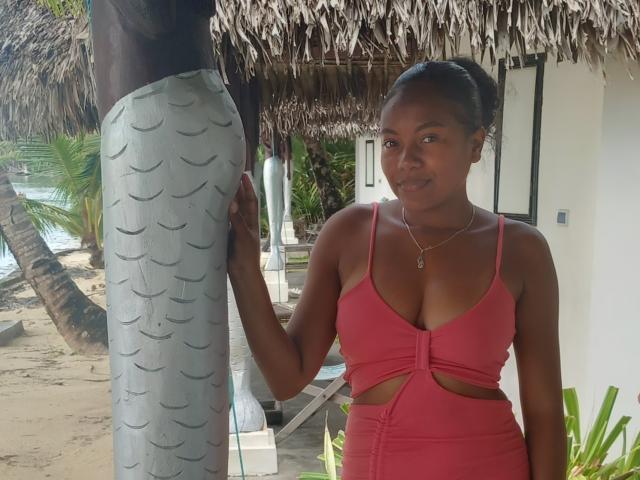 MiaTight with dark skin, small breasts, and a pink cutout dress stands next to a man in patterned shorts, touching his thigh. She has a necklace and a slight smile.