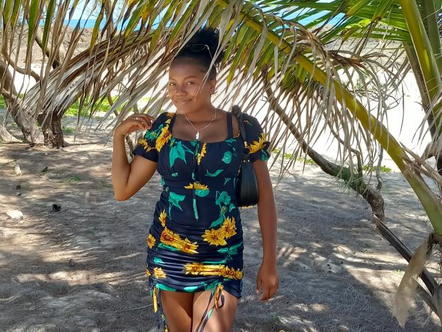 MiaTight with dark skin, black dress, sunflower print, short hair in a bun, black bag, standing under palm leaves, smiling, sunlight on her face, sandy ground.
