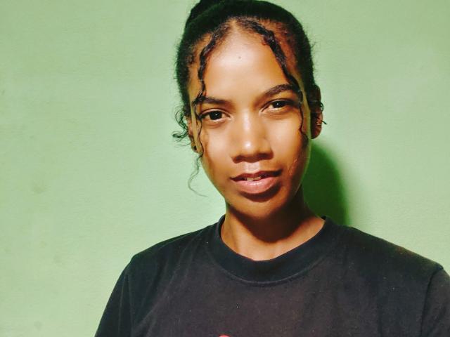 Marrah with dark brown skin, small nose, and medium-sized lips, wearing a black shirt, has short, curly hair, and a serious expression. Green background.