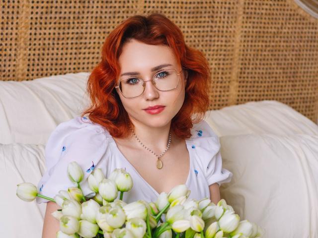 TeresaBirt with red curly hair, glasses, white blouse, holding white tulips, sitting on white couch, wearing a gold necklace, smiling gently. Boobs are not visible.