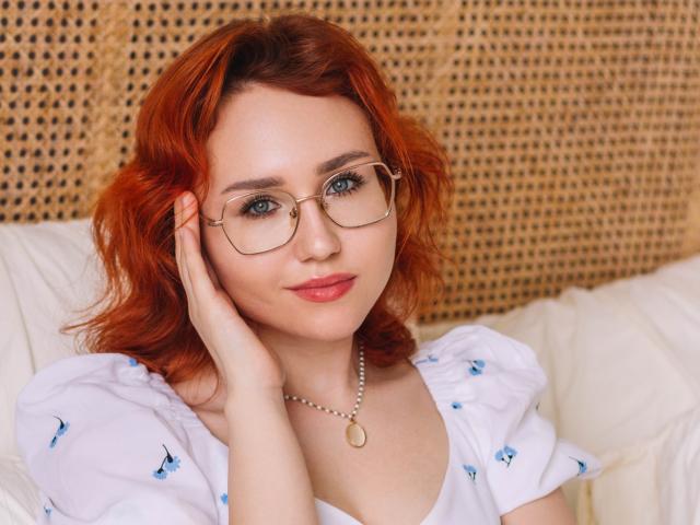 TeresaBirt with red hair, glasses, and white dress with blue butterflies, touching her cheek, wearing a pearl necklace, smiling with pink lipstick, looking cute and sexy.