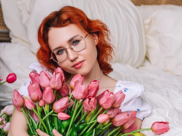 TeresaBirt with red hair, glasses, and white shirt, holding pink tulips, smiling softly, looking at camera, green stems in foreground.