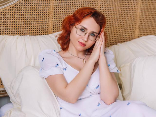 TeresaBirt with red hair, glasses, and white dress with blue butterflies, hands on face, sitting on a wicker chair with white pillows, looking cute and relaxed.