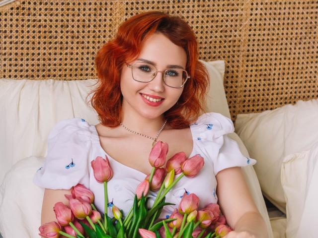 Teresa Birt, com cabelo ruivo, óculos e vestido branco, segurando tulipas rosas, sorrindo, sentada em um sofá de vime, com almofadas brancas, usando um colar, fofa, com os seios visíveis sob o vestido, relaxada, feliz..