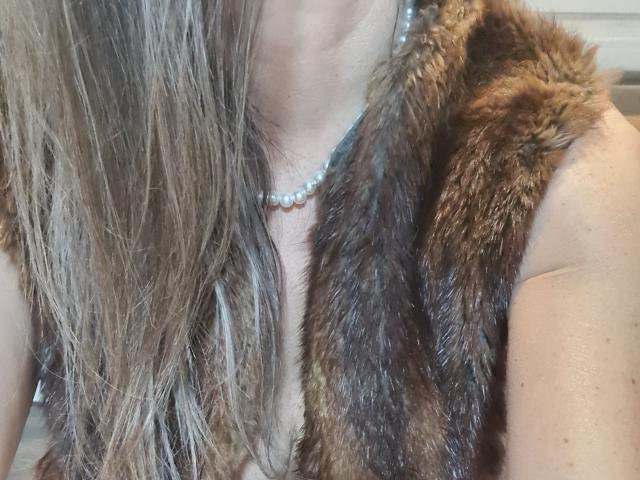 FrenchPlumeX with long, wavy brown hair wearing a brown fur vest and a pearl necklace. Boobs visible, fur vest open. Soft, natural light. Close-up shot.