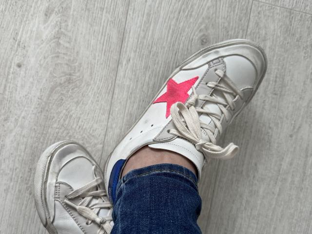 Raichell's white sneakers with a red star on the side, laced up, over blue denim jeans, on a wooden floor. Slightly worn, with loose white shoelaces.
