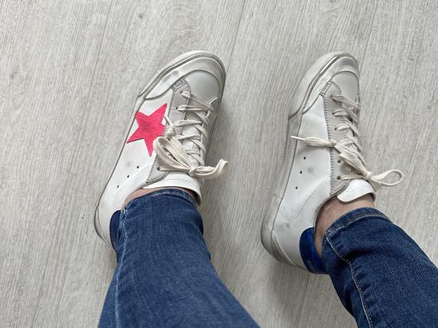 Raichell's white sneakers with pink star on the left shoe, white laces, and blue denim jeans, standing on a light wood floor.
