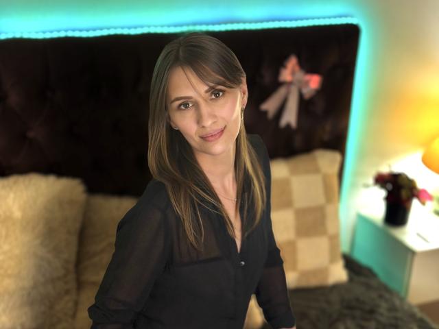 ChristyBrooks, with long brown hair, wearing a sheer black blouse, sits on a bed with a soft smile, showing her tits slightly through the fabric.
