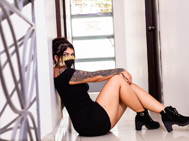 AniisaAli sitting on floor, black dress, black boots, tattooed arm, gold mask, legs crossed, white tile, door, window, black hair in bun, sexy, curvy, tan skin.