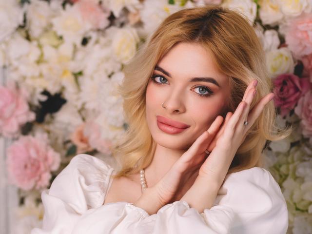 AdeleClifford with blonde hair, blue eyes, and pink lips, wearing a white dress, hands touching her face, pink nail polish, and a pearl necklace, smiling softly.