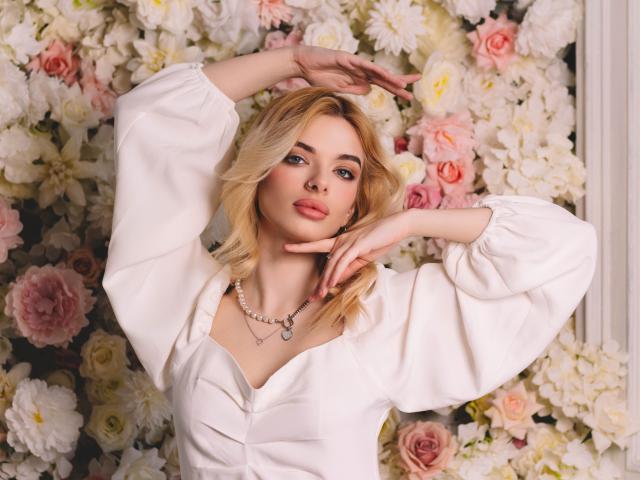 AdeleClifford with blonde hair, wearing a white puffy-sleeve shirt, arms raised, finger on cheek, necklace, pink roses and white flowers in the background, looking at camera.