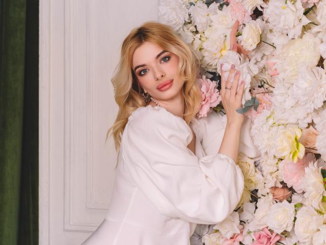 AdeleClifford with blonde hair, blue eyes, and pink lipstick, wearing a white blouse, touches pink and white flowers with her right hand, looking seductive and beautiful.