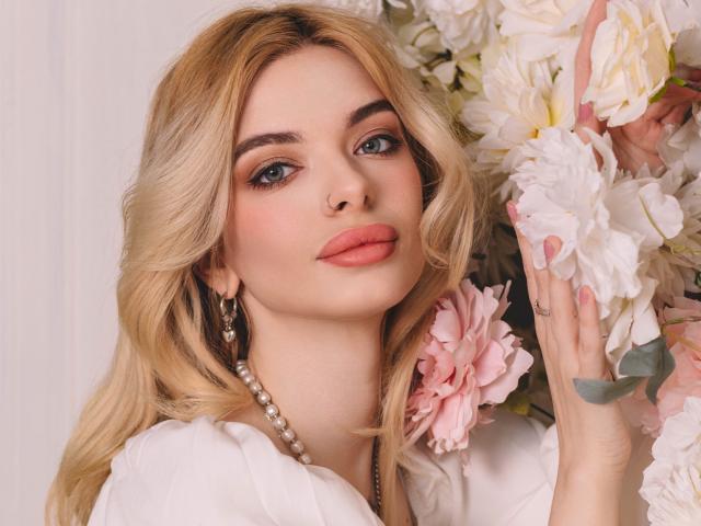 AdeleClifford with blonde hair, blue eyes, and pink lips, holding white and pink flowers, wearing a white shirt and pearl necklace, looking seductive.