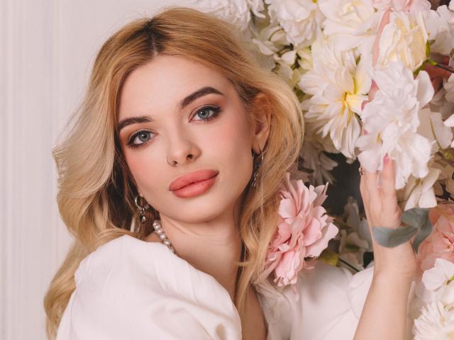 AdeleClifford with blonde hair, pink lipstick, and blue eyes, holding pink flowers, wearing a white shirt and pearl necklace, has a nose ring and a green bracelet.