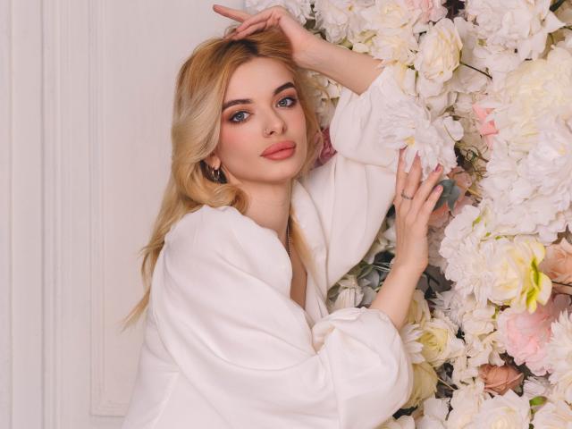AdeleClifford with blonde hair, wearing a white satin robe, touching a wall of fluffy pink and white roses with her right hand, looking at the camera with a slight smile.