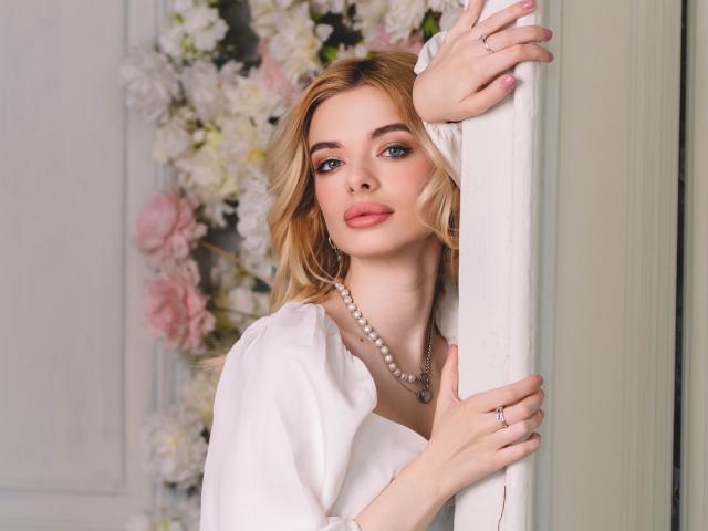 AdeleClifford with blonde hair, wearing a white blouse and pearl necklace, holding a pillar, showing off her tits and pink nail polish, with a ring on her finger.