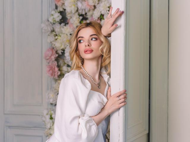 AdeleClifford with blonde hair, wearing a white dress and pearl necklace, leaning against a wall, looking to the side with a slight smile, hand touching the wall.
