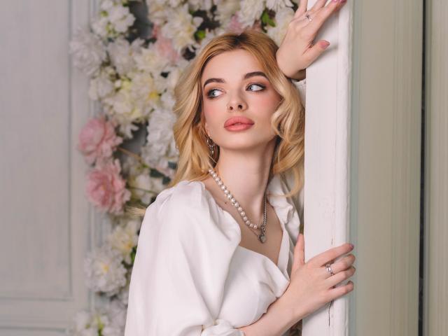 AdeleClifford with blonde hair, white shirt, pearl necklace, and ring, leaning on a white pillar, looking up with pink lips, soft makeup, and elegant, sexy vibe.