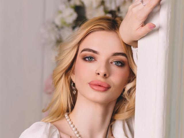 AdeleClifford has blonde hair, blue eyes, pink lips, and is wearing a white dress with a pearl necklace, leaning on a white wall with her hand up.