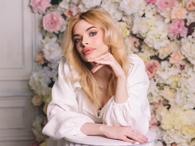 AdeleClifford with blonde hair, blue eyes, and pink lips, wearing a white shirt, resting her chin on her hand, showing off her tits and a cute ring.