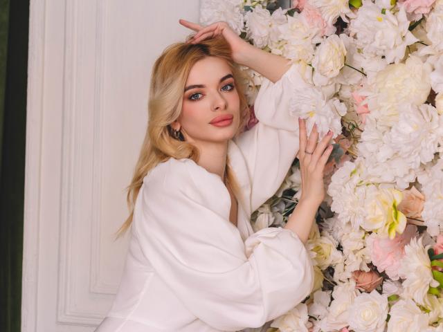 AdeleClifford with blonde hair, white blouse, and pink flowers, touching her head and flowers with one hand, looking at the camera with a subtle smile.